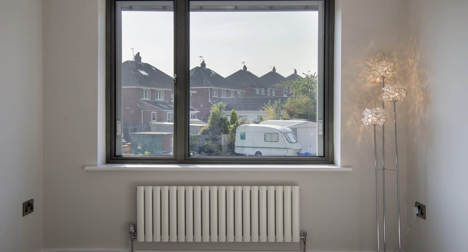 A bright room with a large window featuring bespoke glass solutions, offering views of houses, trees, and a white caravan. Inside, a radiator sits below the window and a modern floor lamp with decorative glass shades stands on the right.