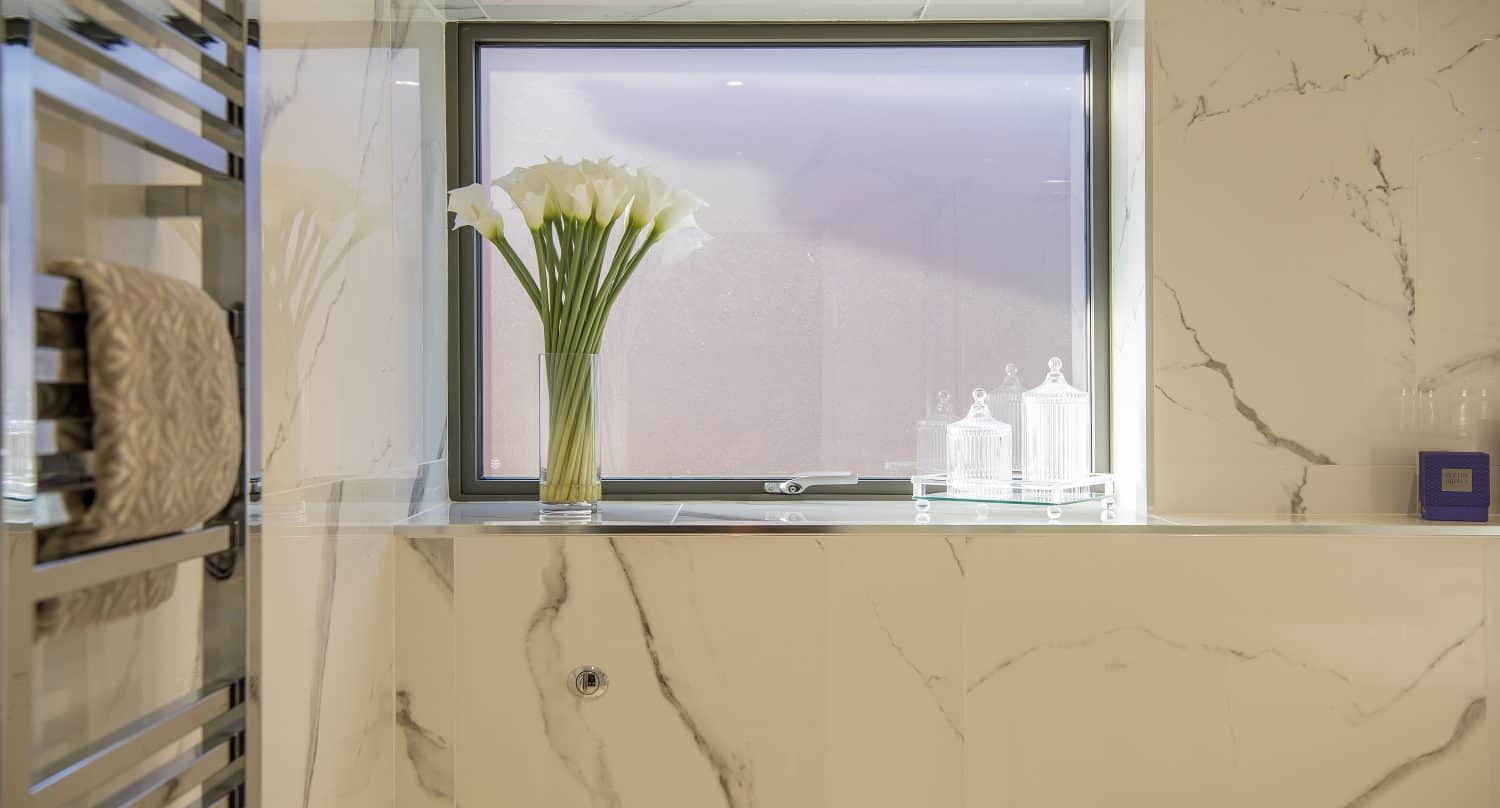 A modern bathroom with marble walls, a window featuring bespoke glass solutions, a vase of white calla lilies on a glass shelf, two glass jars, a purple box, and a metal towel rack with a folded towel.