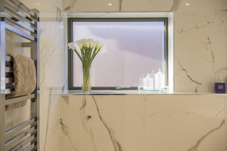 A modern bathroom with marble walls, a window featuring bespoke glass solutions, a vase of white calla lilies on a glass shelf, two glass jars, a purple box, and a metal towel rack with a folded towel.