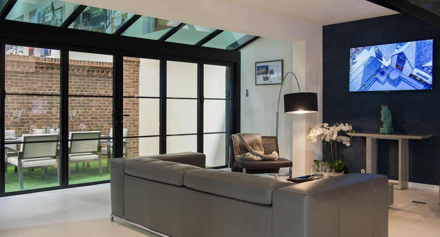 Modern living room with a gray sofa, armchair, floor lamp, and wall-mounted TV. Bespoke glass solutions feature as glass bifold doors lead to a dining area with exposed brick walls and a long table. Neutral tones and contemporary decor.