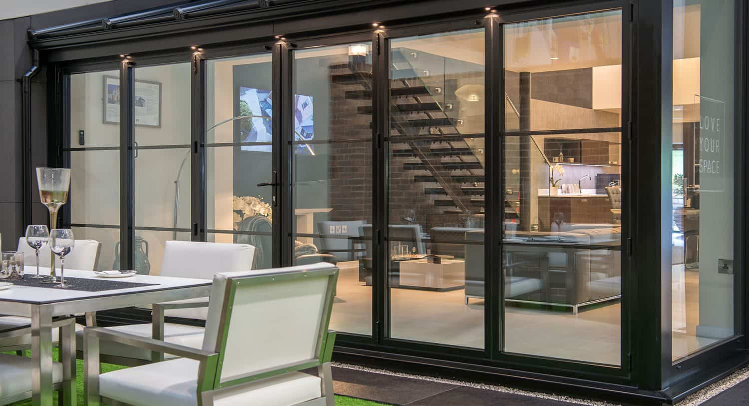 Modern sliding doors open to a stylish, contemporary interior with a staircase, living area, and kitchen. In the foreground, a patio dining set with white chairs and tableware sits on artificial grass. Bespoke glass solutions add elegance throughout.