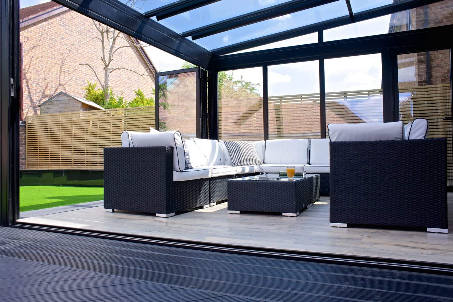Modern patio with black wicker furniture and white cushions under sleek glass roofing, surrounded by glass walls and sliding doors. A low table holds a glass of orange juice. Wooden fence and grassy area are visible outside.