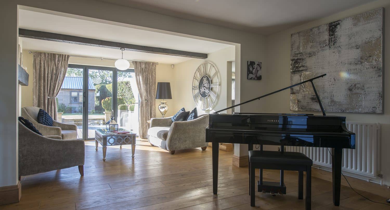 A bright living room with wooden floors, a black grand piano, modern armchairs, a large wall clock, a glass coffee table, and bespoke glass solutions in the floor-to-ceiling windows letting in natural light from the garden.