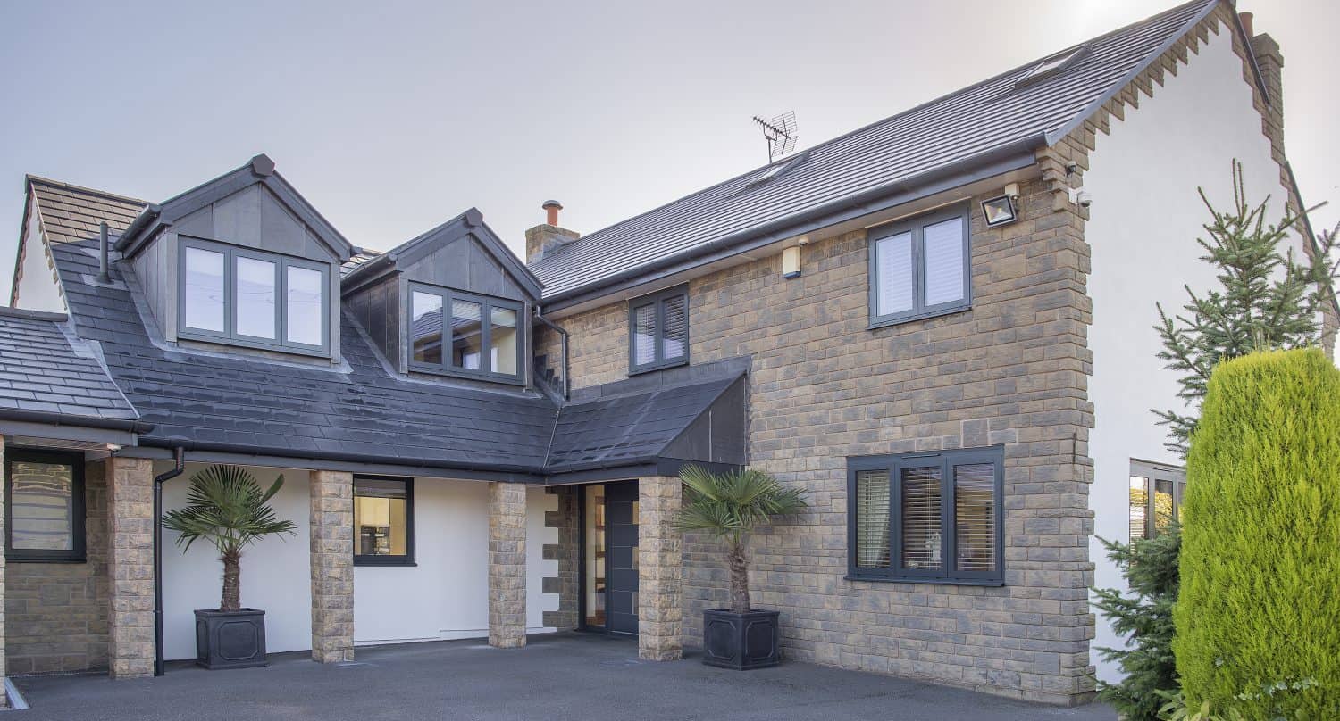 Modern two-story house with a mix of stone and white exterior, dark roof, large windows, and sliding doors. Two potted palm trees frame the entrance. The spacious driveway shines under the sun visible behind the roof.