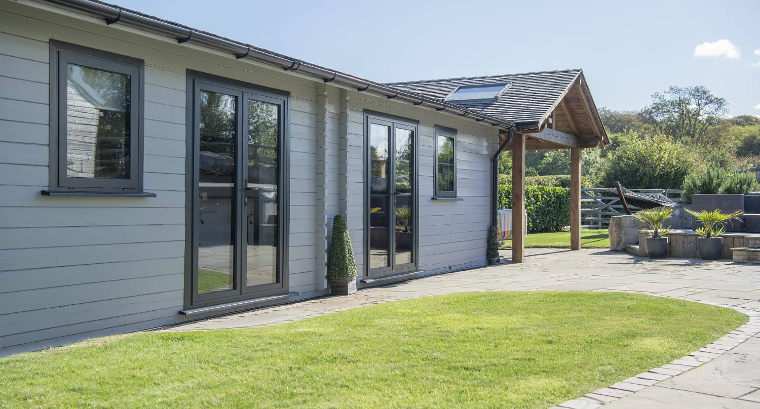 A modern single-story house with light gray siding, large sliding doors and windows featuring bespoke glass solutions, overlooking a curved stone patio and green lawn, with outdoor seating and trees in the background under a sunny sky.