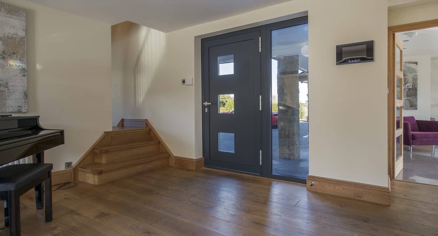 A modern entryway with a dark front door, wooden stairs, and hardwood floors. A black piano sits on the left, while bespoke glass solutions like sliding doors reveal a purple sofa in the adjoining room.