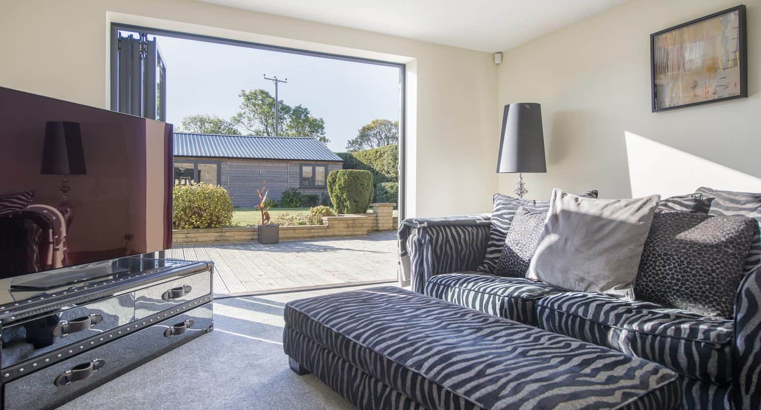 Bright living room with a zebra-patterned sofa and ottoman, a glass TV stand with a large TV, and sliding doors leading to a sunny patio and garden with shrubs and a small building outside.