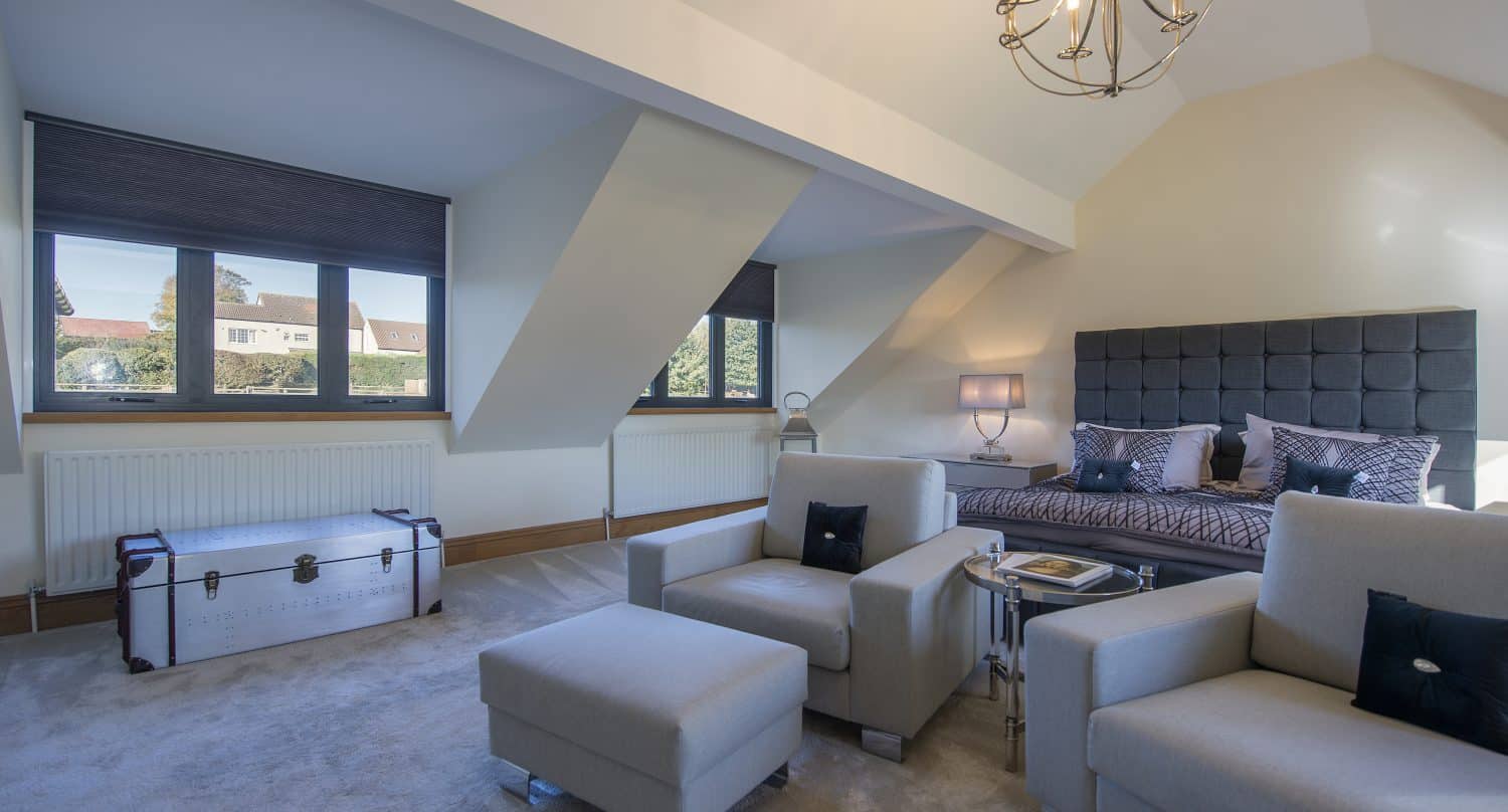 A spacious, modern bedroom with large windows, a dark upholstered bed, two gray armchairs, a matching ottoman, a white trunk, and a chandelier—neutral tones and bespoke glass solutions fill the room with abundant natural light.