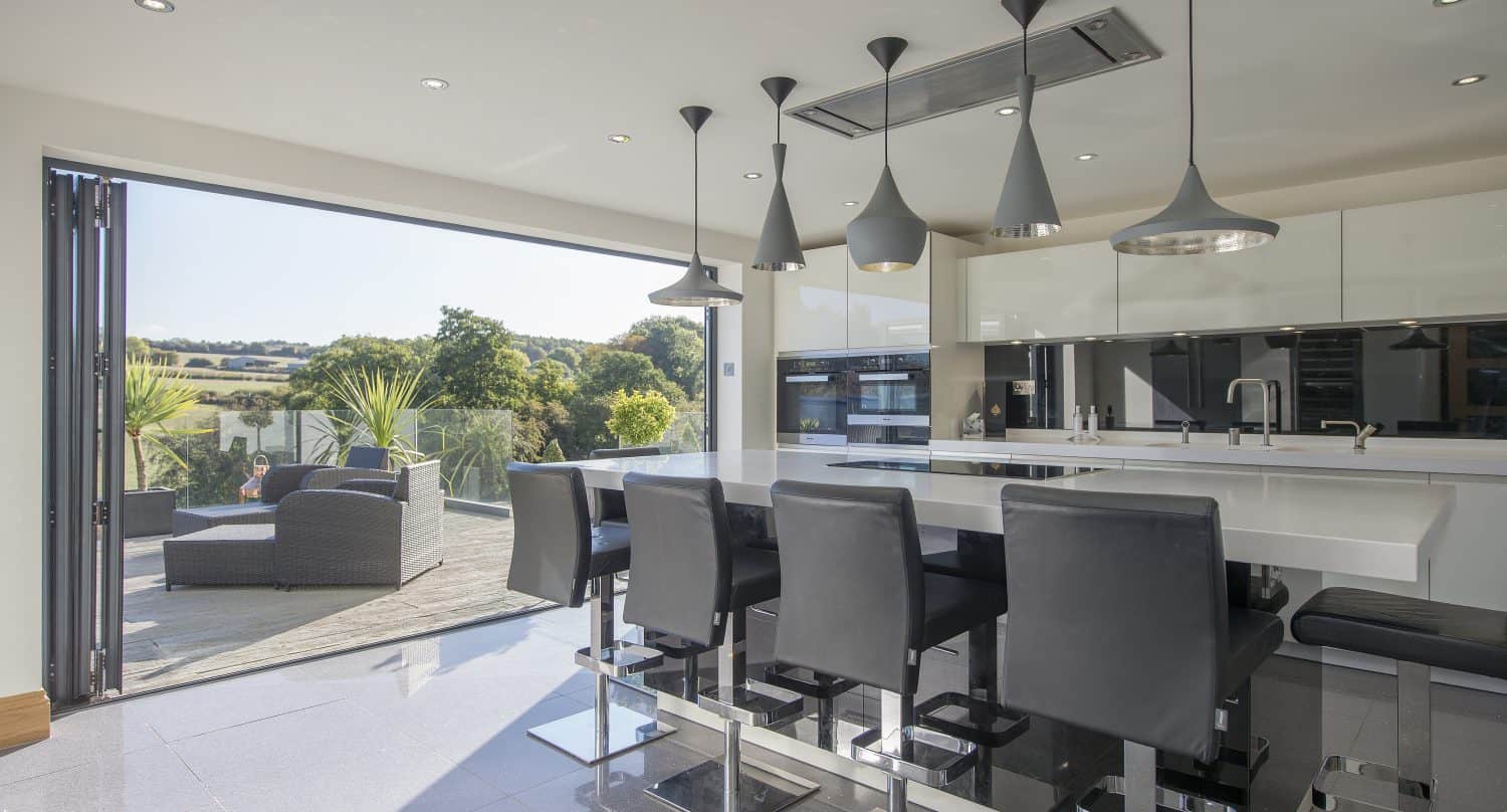 Modern kitchen with black chairs at a white island, pendant lights above, and bespoke glass solutions like bifold doors open to a patio with outdoor seating, overlooking green trees and a scenic landscape. Bright, airy, and contemporary design.