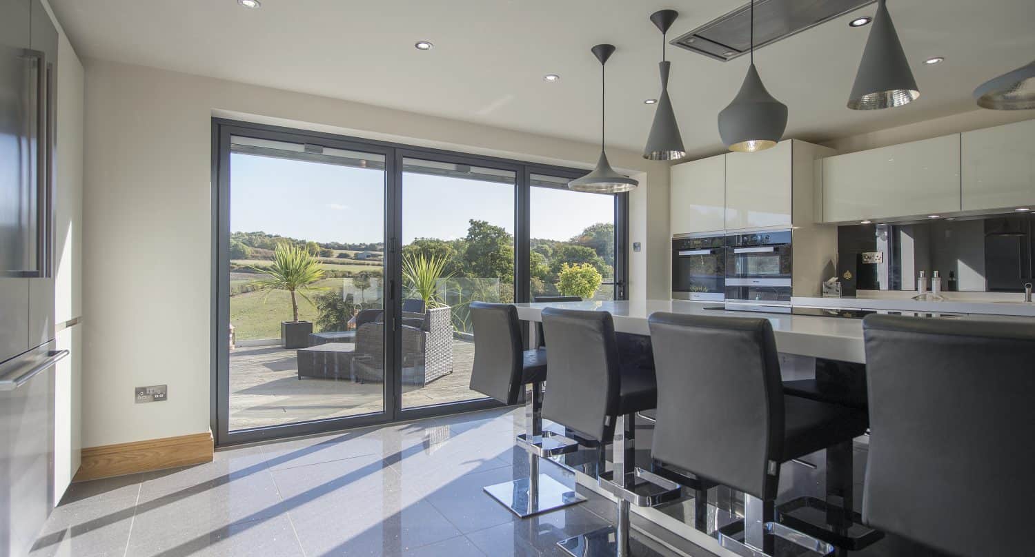 Modern kitchen with black chairs around a white island, pendant lights above, and large sliding doors opening to a patio with outdoor furniture overlooking a scenic, grassy landscape. Sunlight streams into the room.