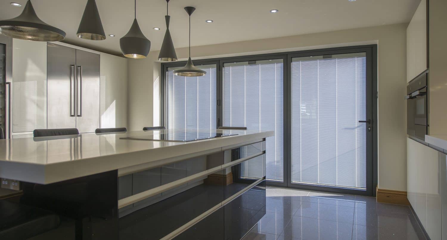 Modern kitchen with glossy black cabinets, a large white island, pendant lights, built-in appliances, and bifold doors letting in natural light. The space appears sleek and minimalistic with bespoke glass solutions enhancing its style.