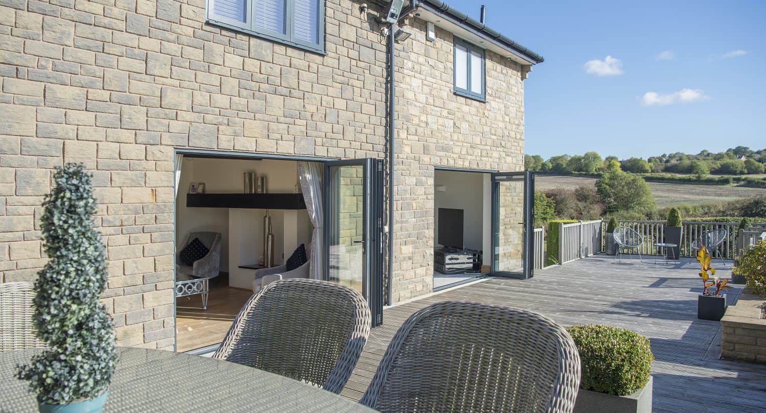 A modern stone house with bespoke glass solutions features large sliding glass doors opening onto a wooden deck with outdoor furniture. The deck overlooks a scenic countryside landscape under a clear blue sky.