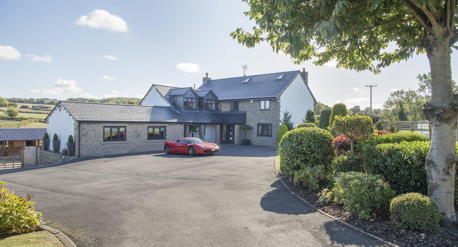 A large, modern house with stone walls and dark roof tiles, featuring sleek sliding doors, is surrounded by greenery and gardens. A red sports car is parked on the spacious driveway under a sunny, blue sky.