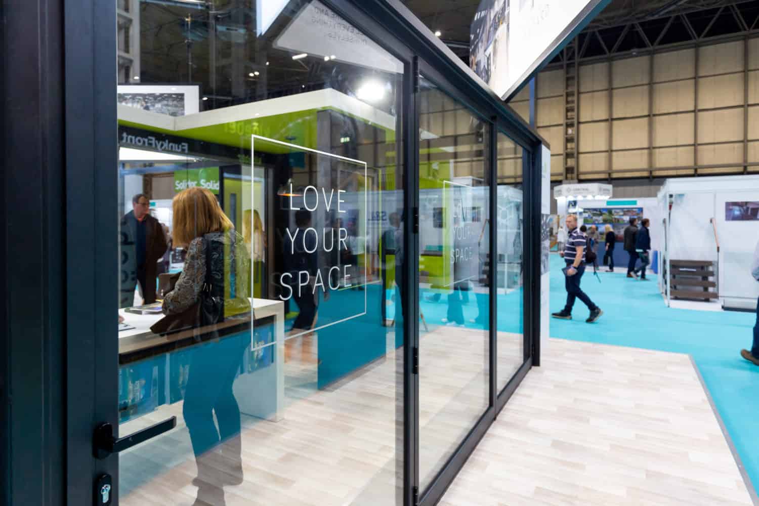 A glass-walled booth at an exhibition features the words LOVE YOUR SPACE printed on the glass. Showcasing bespoke glass solutions, people are walking and interacting inside the booth and around the exhibition hall.