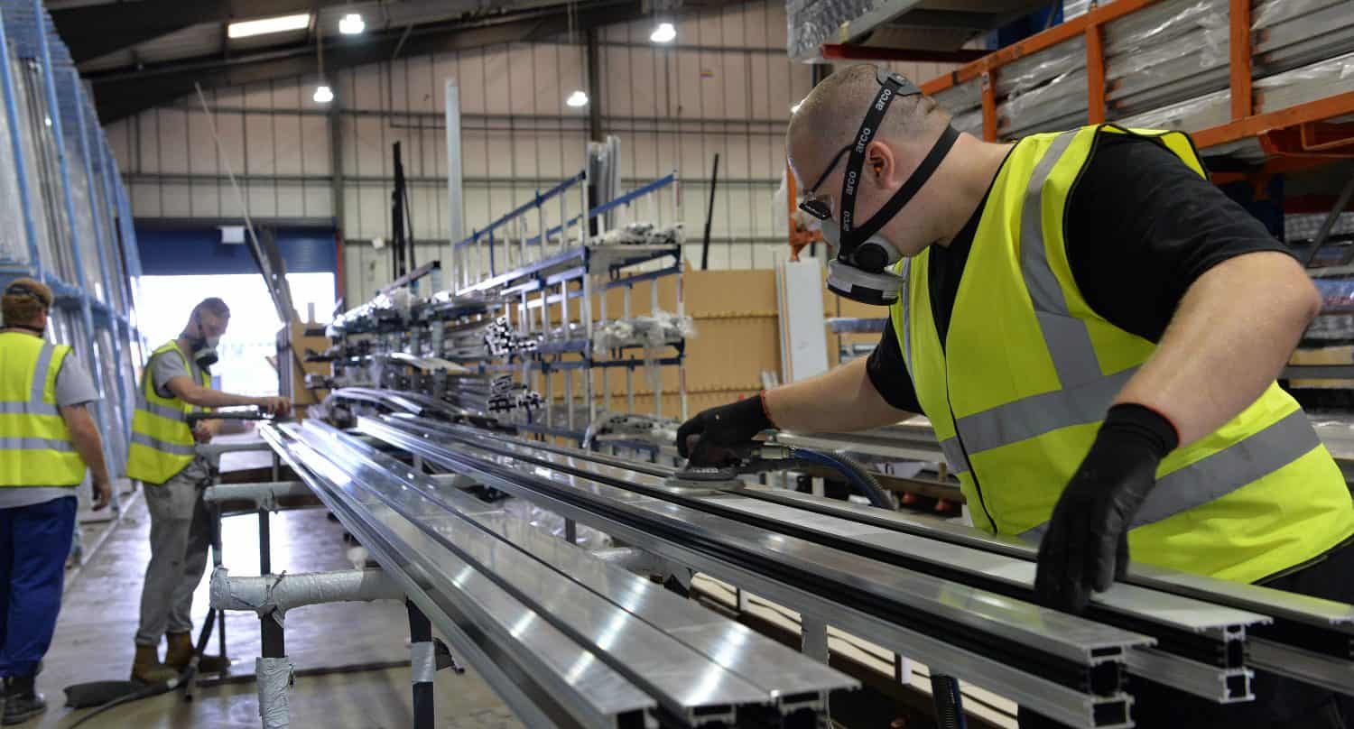 A worker in a high-visibility vest and mask handles metal beams in a large industrial warehouse with glass roofing, while another worker in the background also works with materials on long racks.
