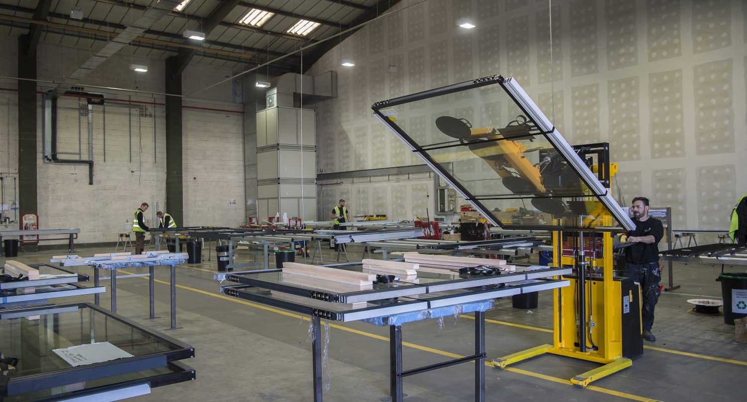 Workers in a large factory space use machinery to handle large glass panels, including those for sliding doors. Several glass sheets rest on tables around the area, and high ceilings with bright lights illuminate the industrial workspace.