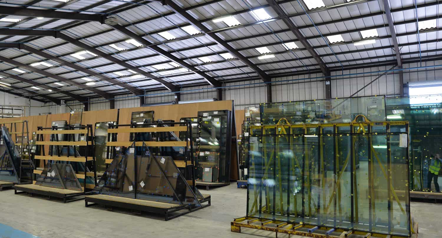 Large industrial warehouse interior with metal beams and a high ceiling, storing numerous large glass panels for bifold doors on metal racks and wooden stands. The space is well-lit with overhead lights.