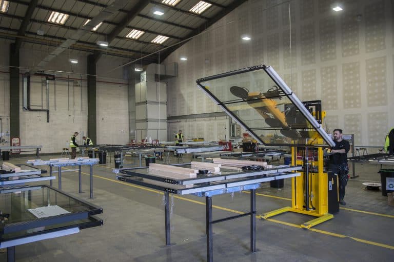 Workers in a large factory space use machinery to handle large glass panels, including those for sliding doors. Several glass sheets rest on tables around the area, and high ceilings with bright lights illuminate the industrial workspace.