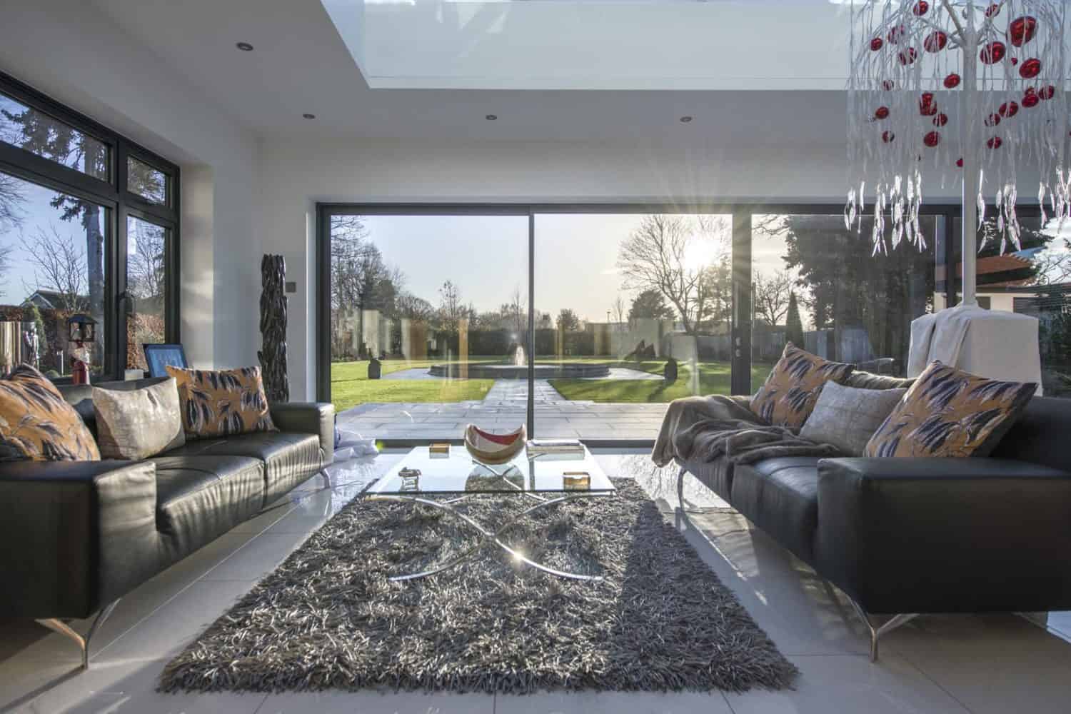 Modern living room with two dark sofas, a glass coffee table, and a gray shag rug. Bespoke glass solutions include large sliding doors that open to a sunny garden and pool. A contemporary chandelier hangs from the ceiling.