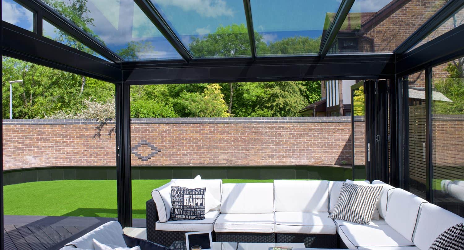 A modern glass conservatory with a white L-shaped sofa, throw pillows, and a coffee table sits beside a green lawn. Large sliding doors and a glass roofing reveal a brick wall and lush trees outside under a blue sky.