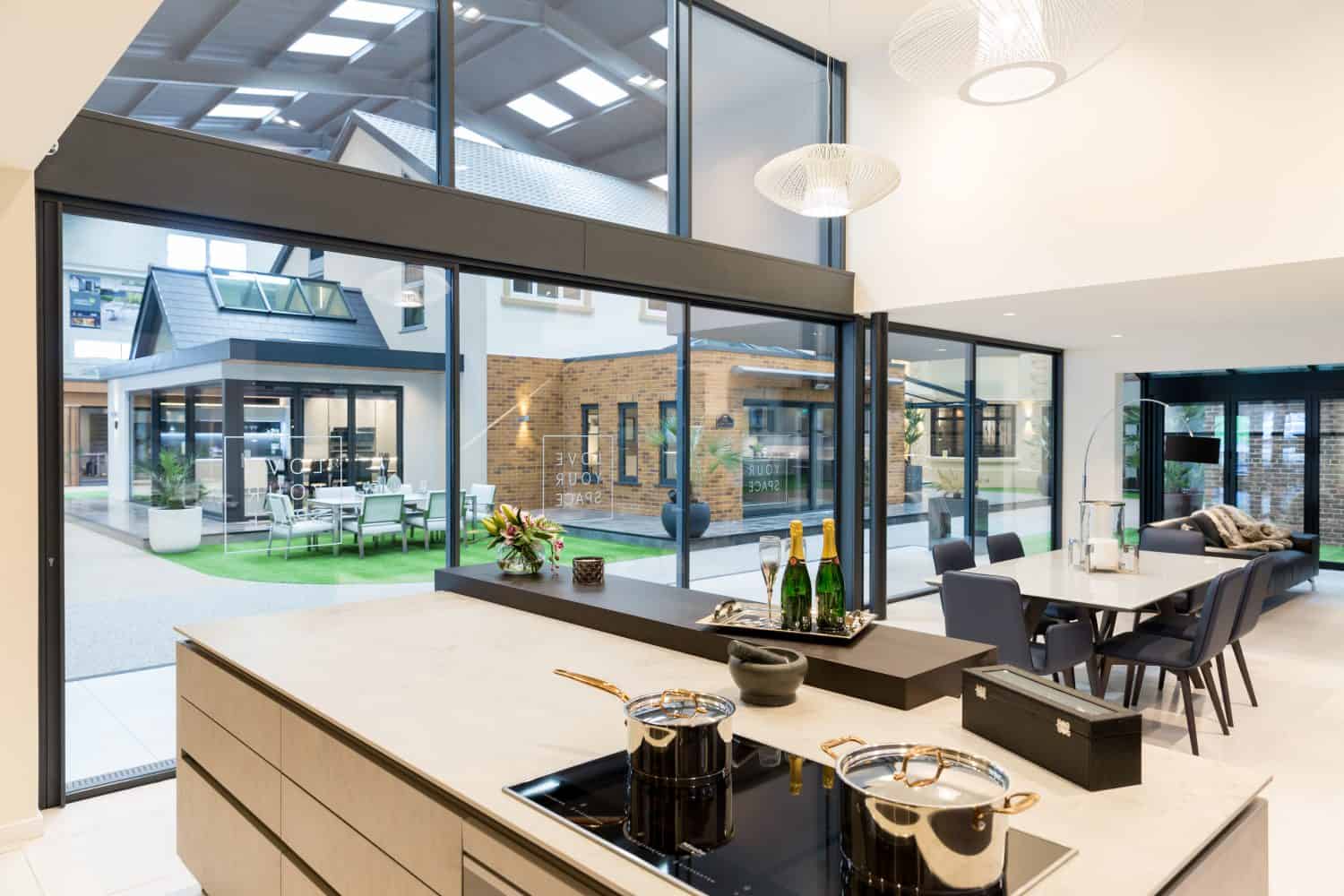 Modern kitchen and dining area with large windows and sliding doors overlooking a bright courtyard and neighboring buildings. The space features a sleek island, induction cooktop with pots, and elegant table settings.