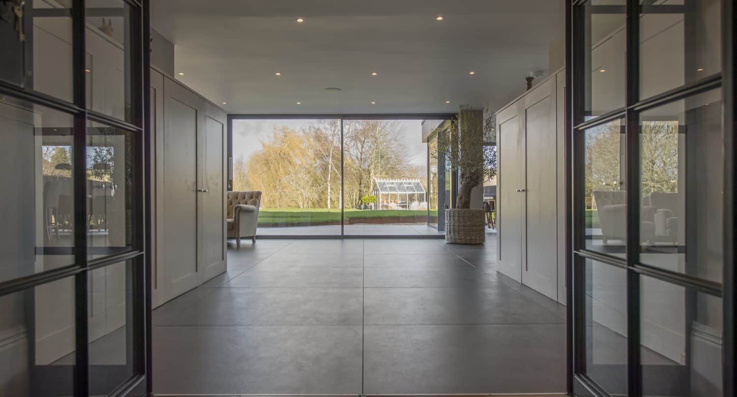 Modern interior hallway with bifold doors leads to a spacious living area. Large floor-to-ceiling windows offer views of a patio and green lawn, with trees and a conservatory featuring glass roofing in the background.