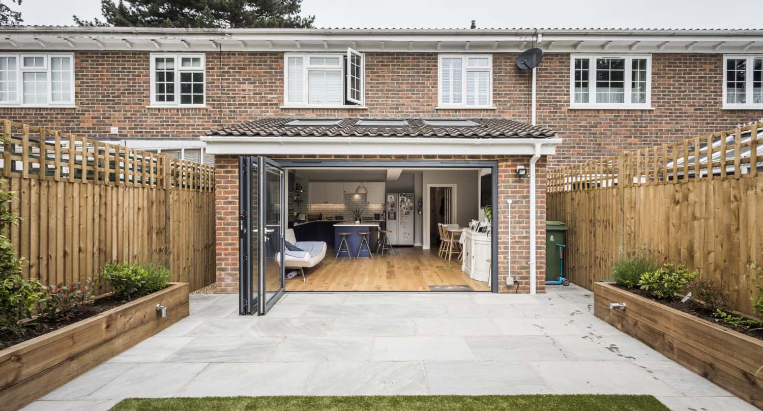 A view of a modern brick house with bifold doors opening to a patio and small backyard, featuring fenced sides, raised garden beds, and a cozy, open-plan kitchen and living area visible inside.