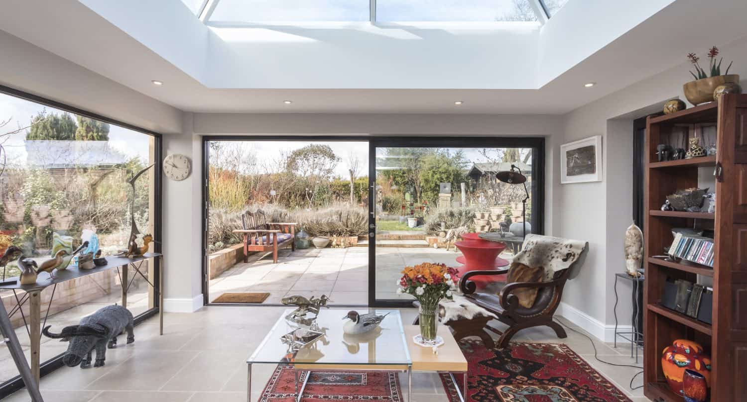 A bright sunroom with large sliding doors, a skylight, eclectic decor, indoor plants, a chair, bookshelves, and a coffee table with flowers opens to a garden patio with outdoor furniture.