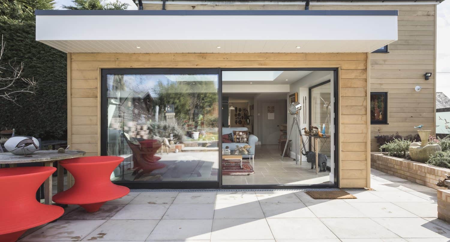 Modern house extension with large bifold doors opening onto a patio with white tiles. Red sculptural outdoor chairs sit on the left; the interior is visible through the glass, showing a cozy living space.