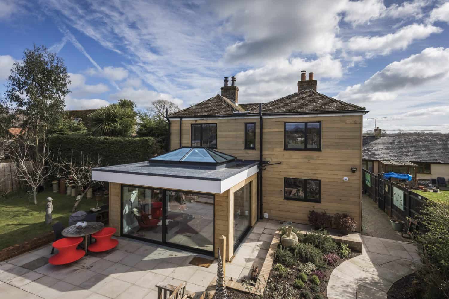 Modern house with tan wooden siding and black-framed windows, featuring a large glass extension with skylight and sleek sliding doors. The patio has red furniture, landscaped garden beds, and is surrounded by trees under a partly cloudy sky.