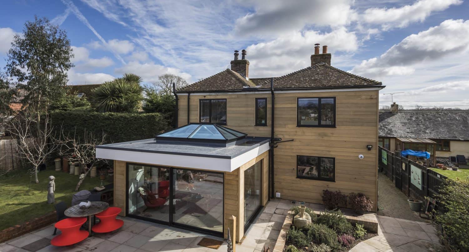 Modern house with tan wooden siding and black-framed windows, featuring a large glass extension with skylight and sleek sliding doors. The patio has red furniture, landscaped garden beds, and is surrounded by trees under a partly cloudy sky.