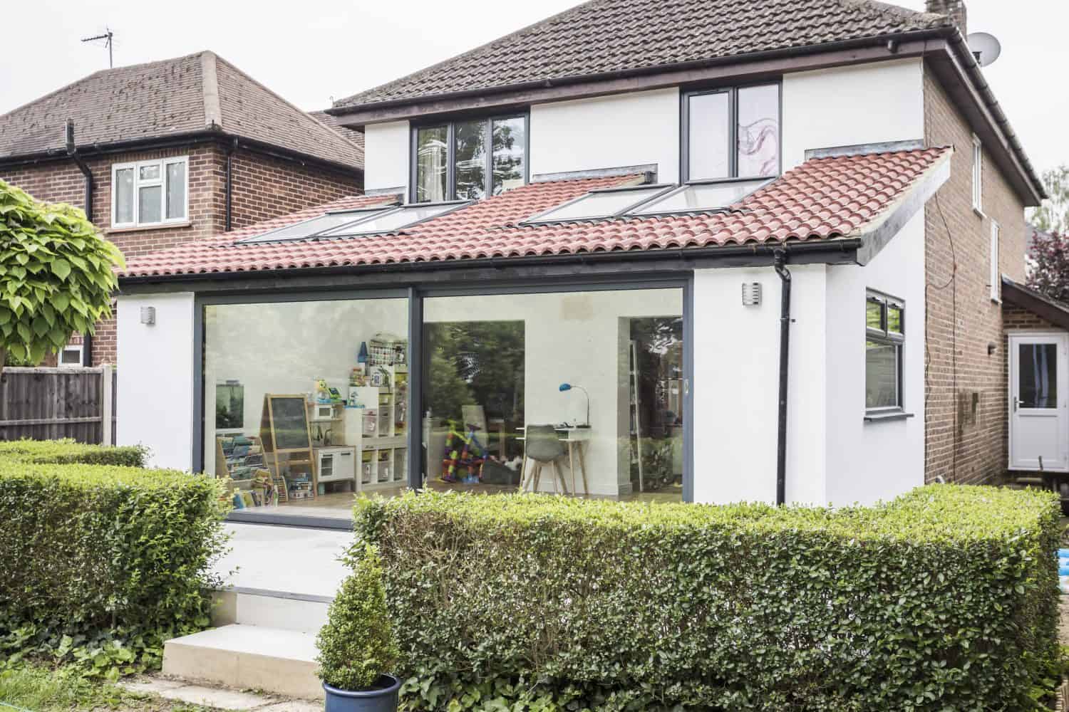 A modern house with a red-tiled roof and bespoke glass solutions, featuring large glass sliding doors opening onto a patio. Inside, childrens toys and books are visible. The garden has neatly trimmed hedges and a potted plant.