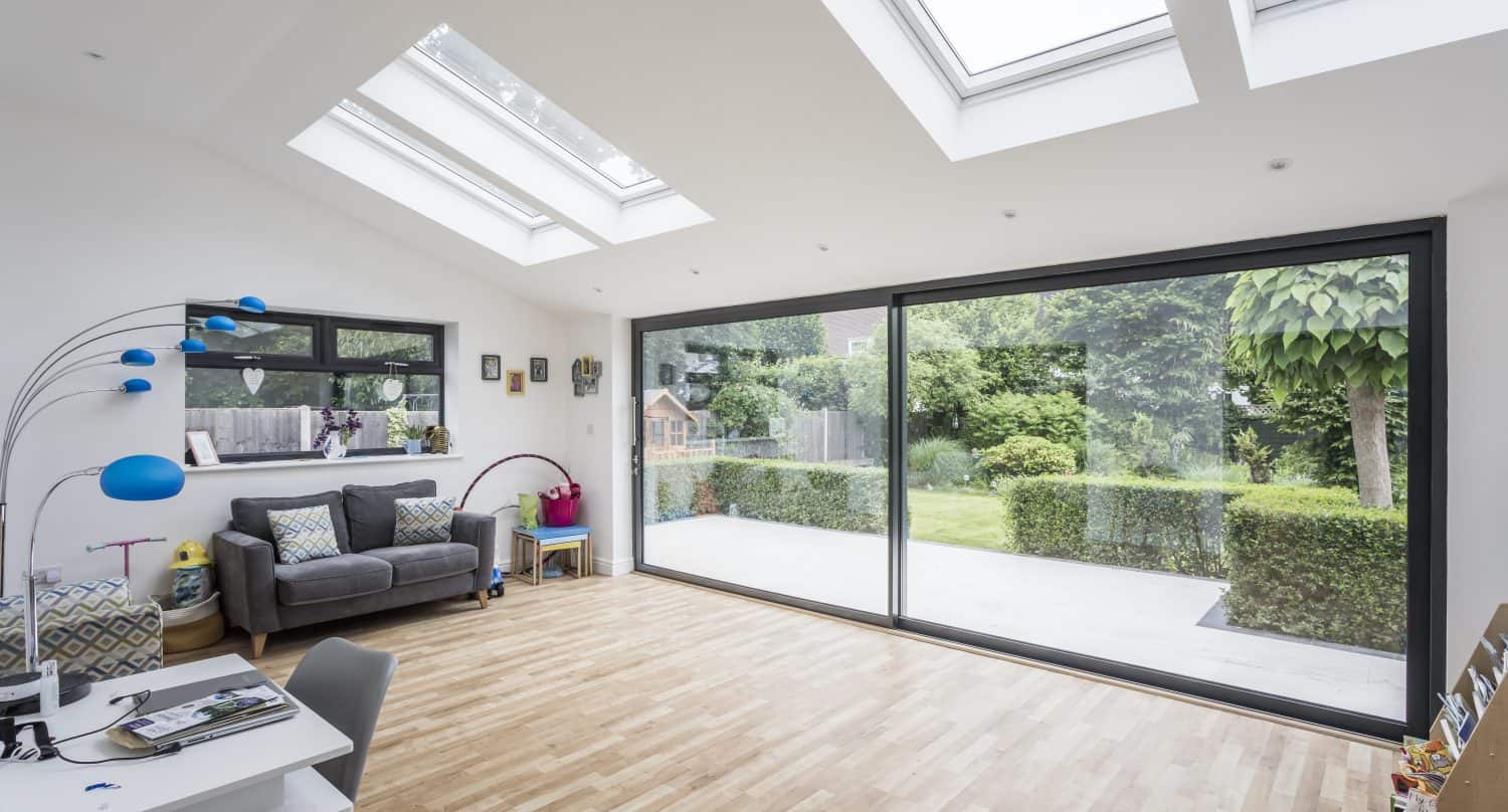 Bright, modern room with wood flooring, skylights, large bifold doors opening to a green garden, a gray sofa, colorful floor lamp, and desk with chair in the corner.