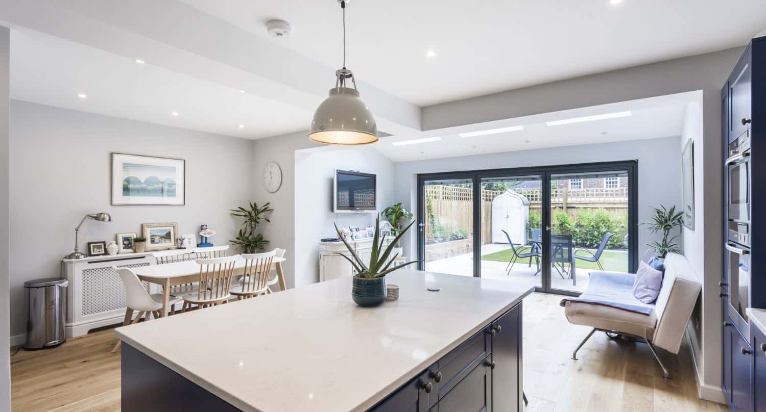 Modern open-plan kitchen and dining area with a large island, wooden floors, stylish decor, and bespoke glass solutions—such as sliding doors—leading to a patio and garden. Natural light fills the bright, spacious room.