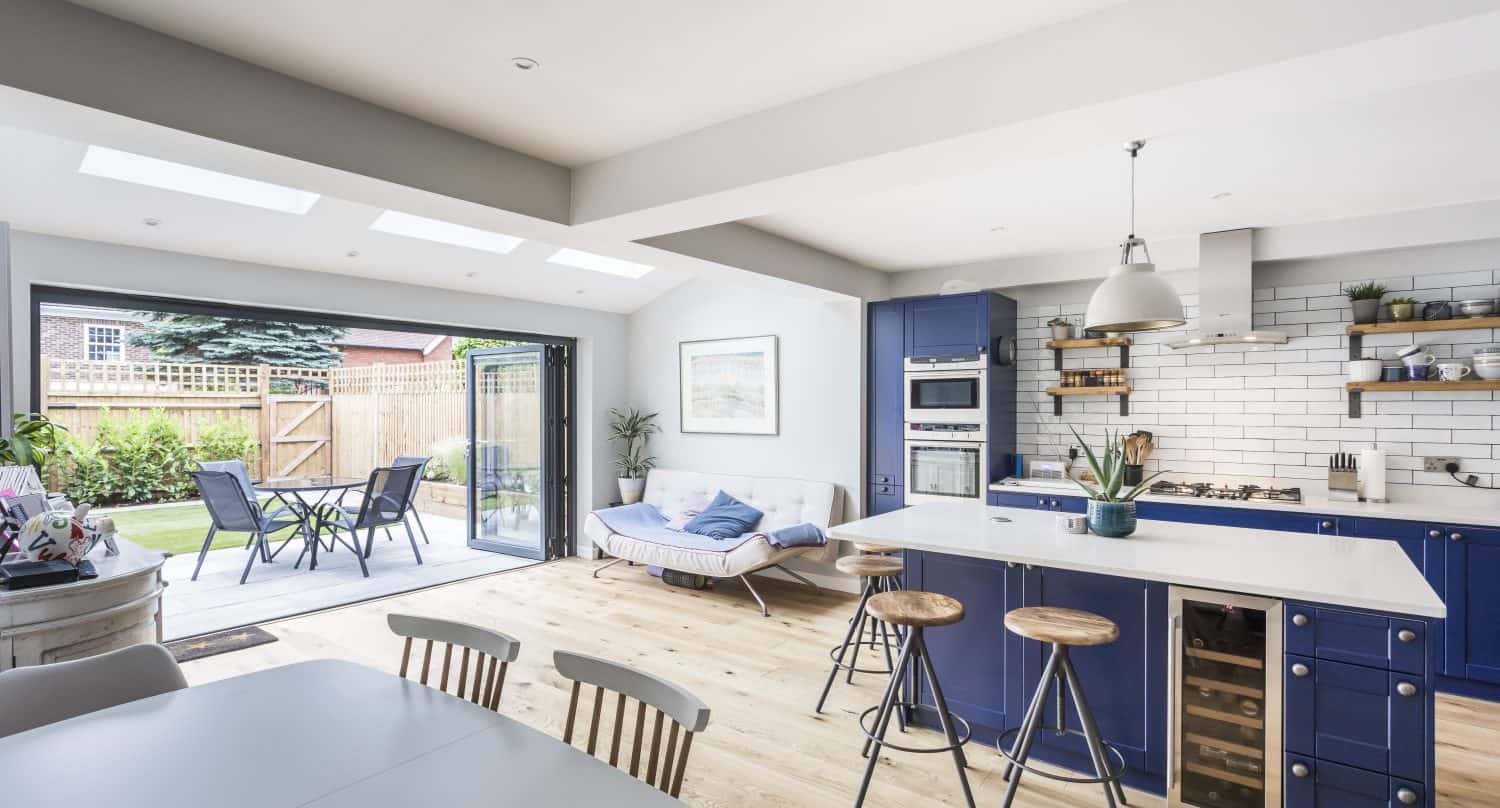 Bright, modern kitchen and dining area with blue cabinets, wooden floor, white island with stools, open shelves, and sliding doors leading to a patio with outdoor seating and a green garden.