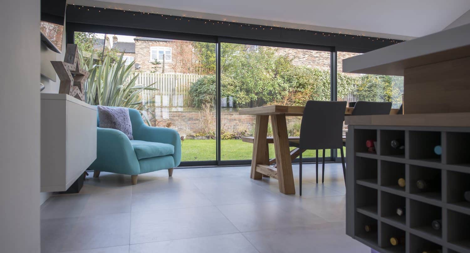 Modern dining area with a large sliding glass door opening to a garden, featuring a turquoise armchair, wooden dining table with black chairs, and a built-in wine rack in the foreground—perfect for showcasing bespoke glass solutions.