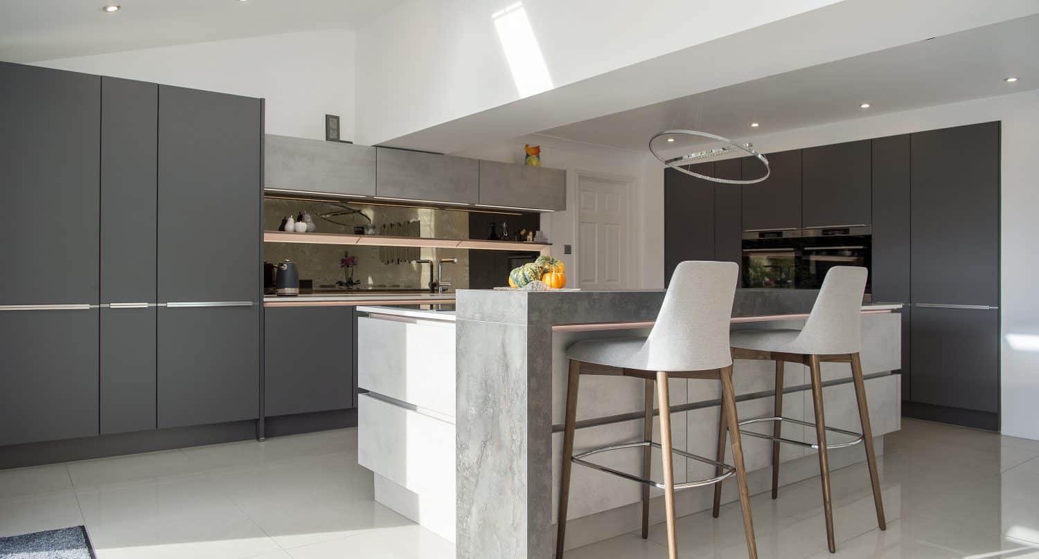 Modern kitchen with sleek dark gray cabinets, a large island with a gray and white finish, two tall bar stools, built-in ovens, and under-cabinet lighting. Sunlight streams in through a skylight and bespoke glass solutions add an elegant touch.