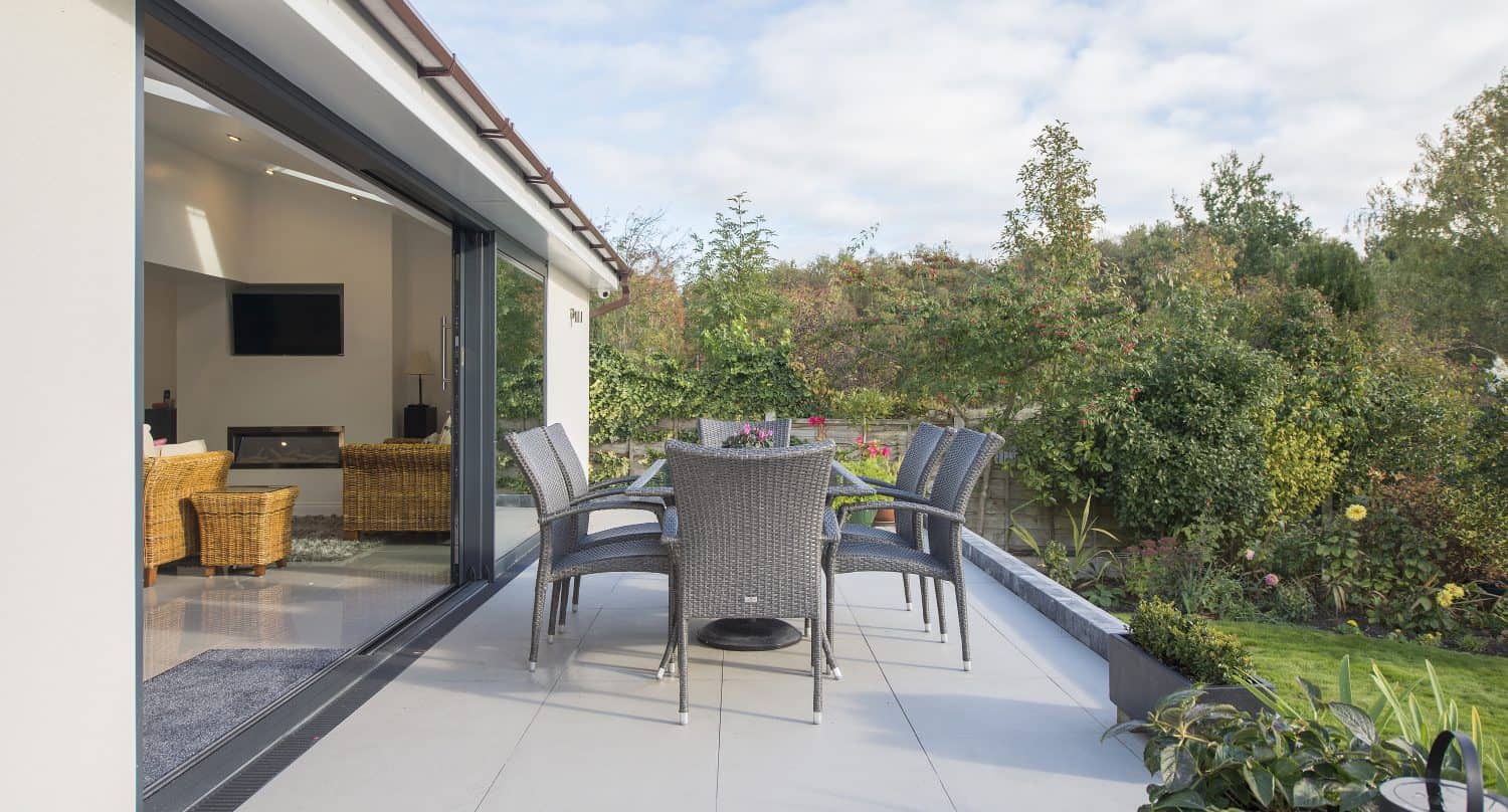 A modern patio with a glass table and six wicker chairs overlooks a lush green garden. Large sliding doors connect the patio to a bright, cozy indoor living area with more wicker furniture.