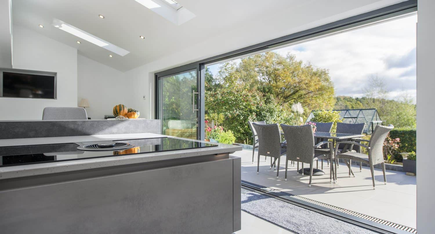Modern kitchen with a sleek countertop opens to a sunny patio through large sliding doors. Outdoor dining table and chairs sit on the terrace beneath bespoke glass solutions, surrounded by greenery and trees under a bright sky.