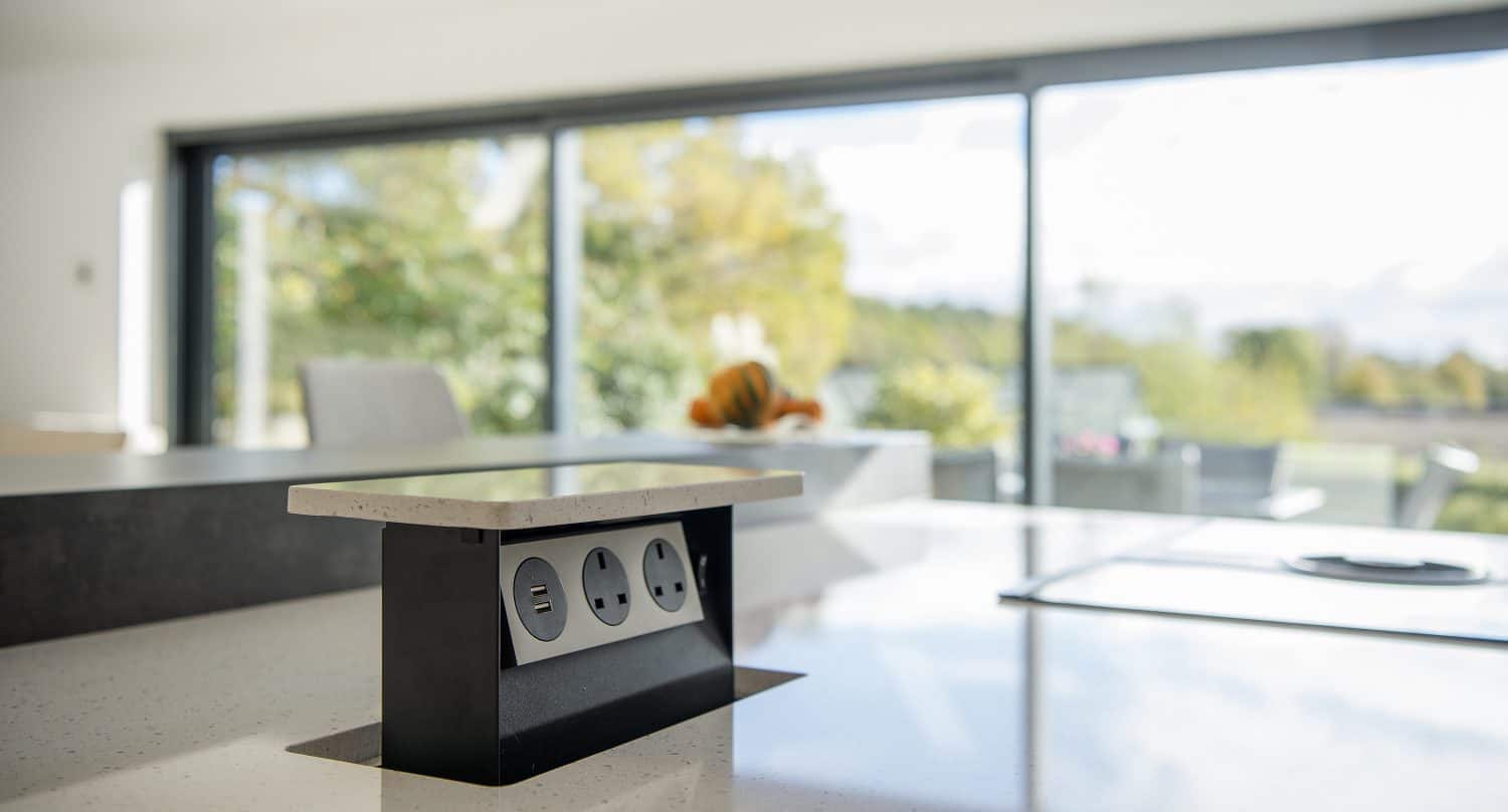 A modern kitchen countertop with a pop-up power outlet featuring USB and plug sockets, set against large windows and sliding doors overlooking a bright, scenic outdoor area.