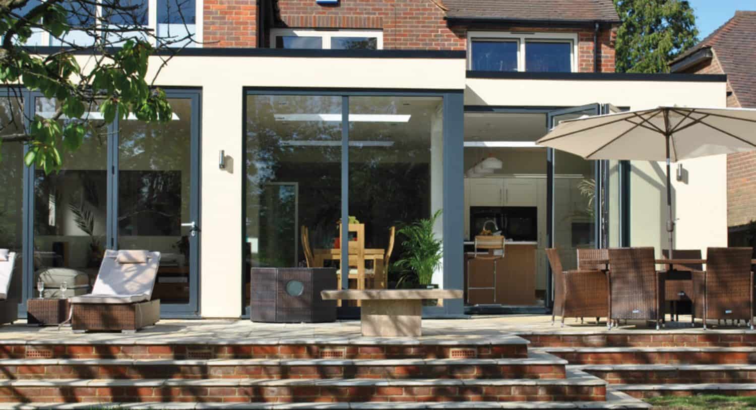 Modern house with large glass sliding doors opening to a patio. The patio features wicker furniture, a dining table, chairs, an umbrella, and a small stone table, with brick steps leading to a garden area and sleek glass roofing overhead.
