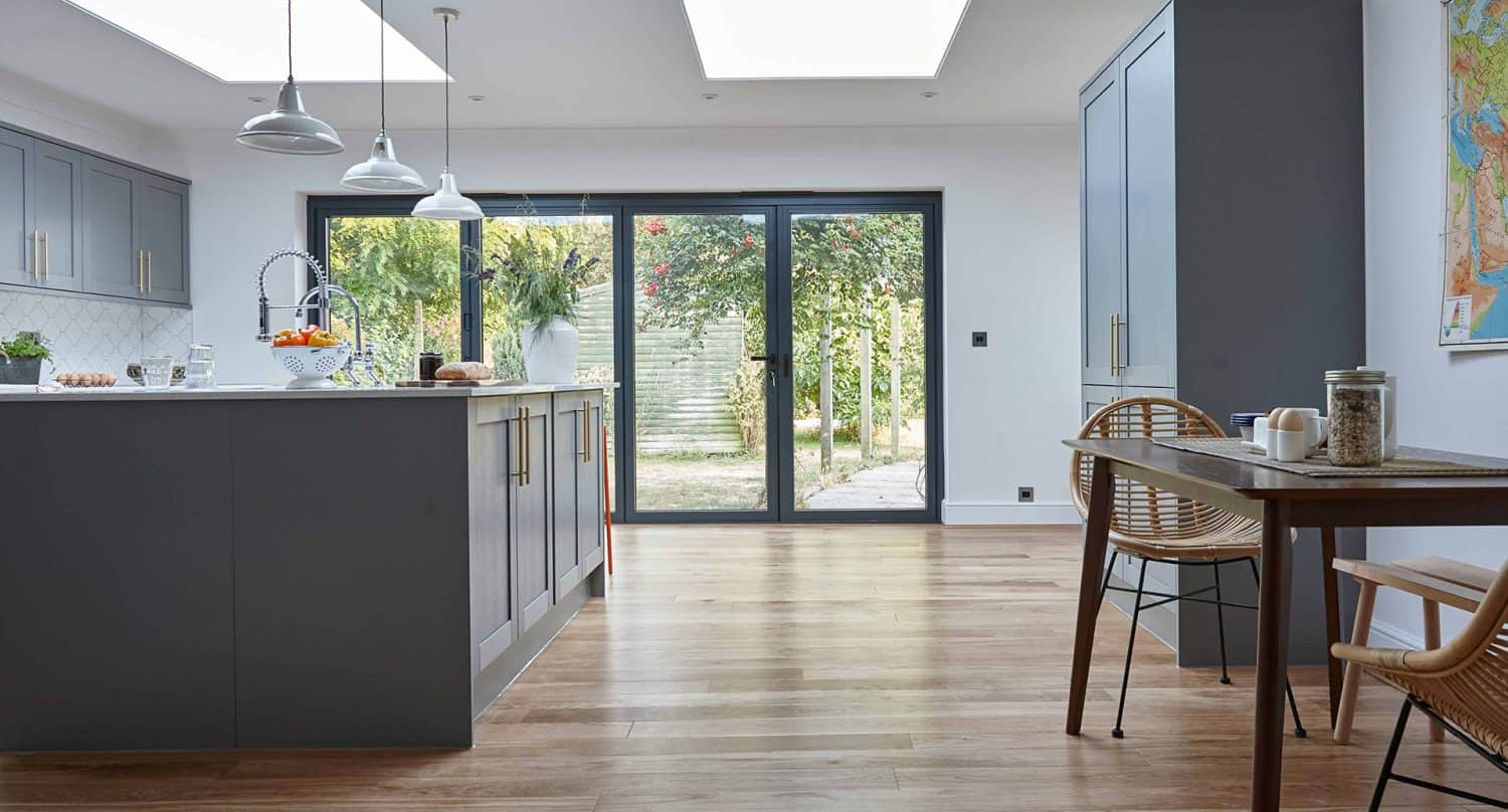 Modern kitchen and dining area with gray cabinets, island, wooden floor, and bifold doors opening to a garden. A wooden table with a chair is on the right, and sunlight streams in through skylights and large doors.