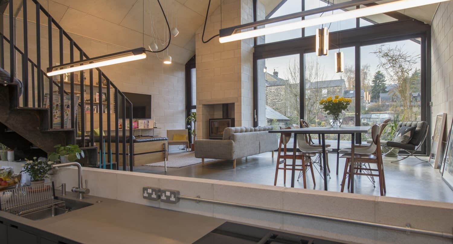 Modern open-plan living space with high ceilings, large windows, and a kitchen in the foreground. Sliding doors open to the outside, while a dining table with flowers and cozy seating area invite relaxation. Natural light fills the room.