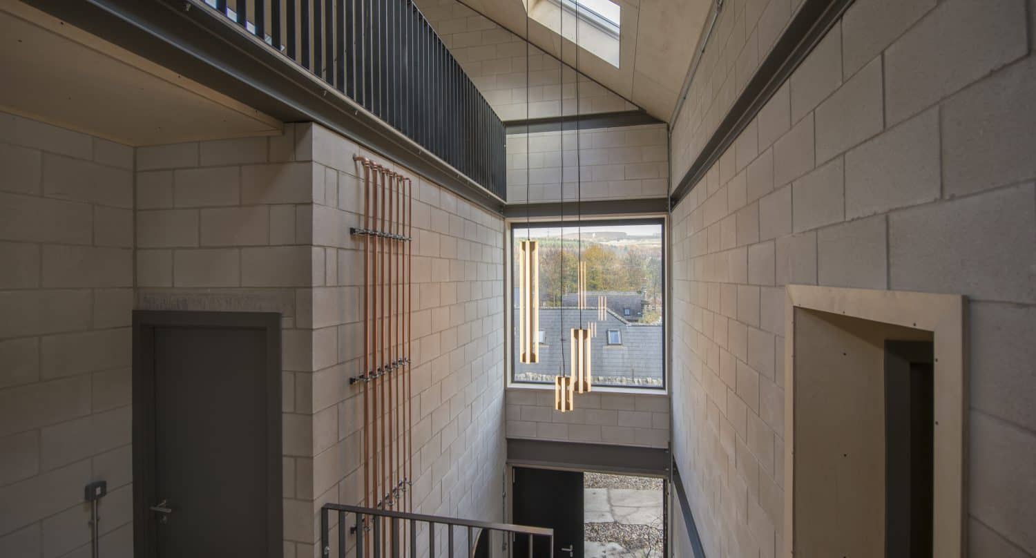 A modern hallway with high ceilings, white cinder block walls, large windows, a skylight, contemporary hanging light fixtures, bespoke glass solutions, and an industrial-style metal railing along the upper floor.