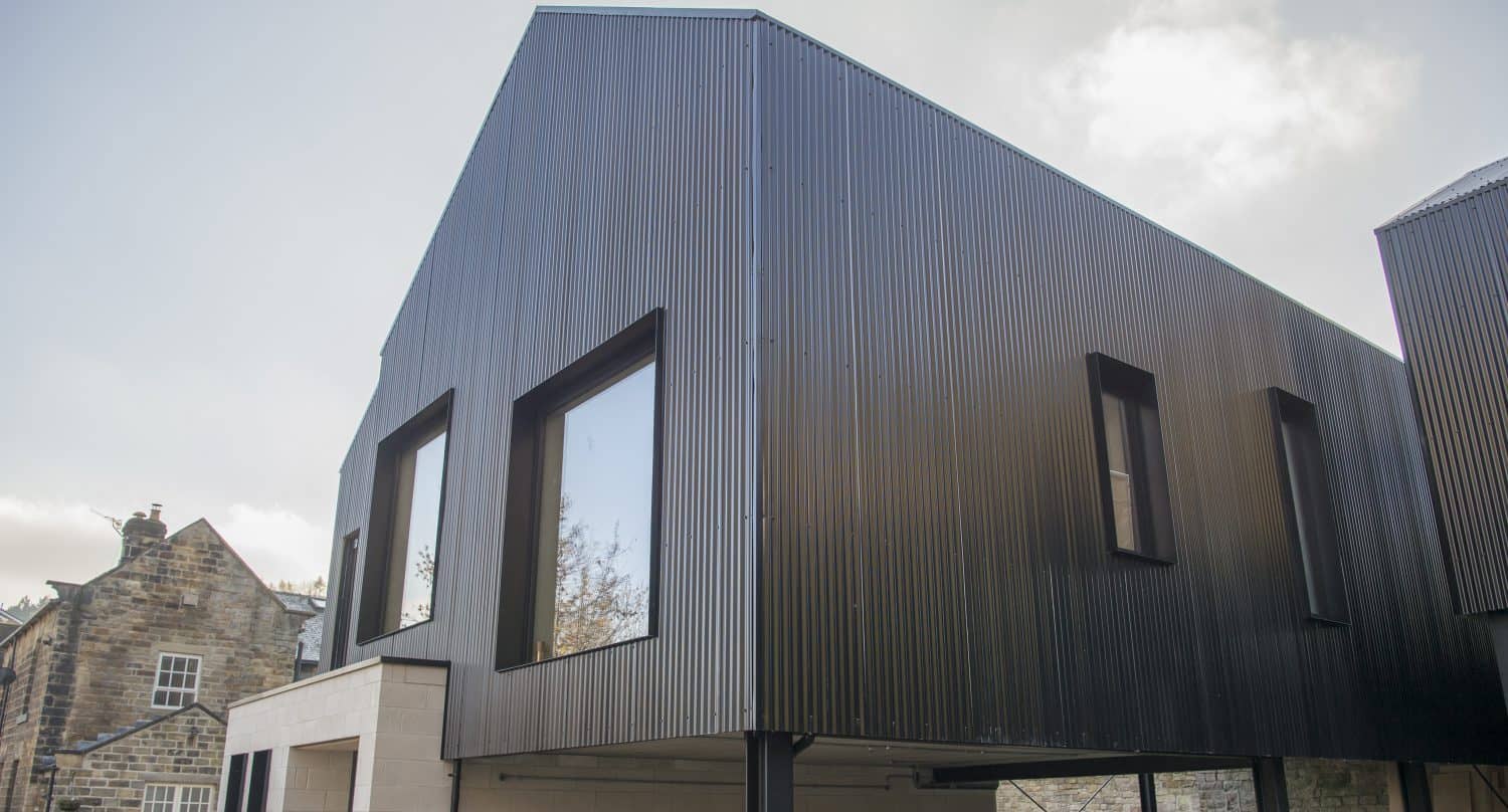 A modern house with black corrugated metal siding, large windows, and sliding doors is elevated on pillars beside a traditional stone building under a cloudy sky.