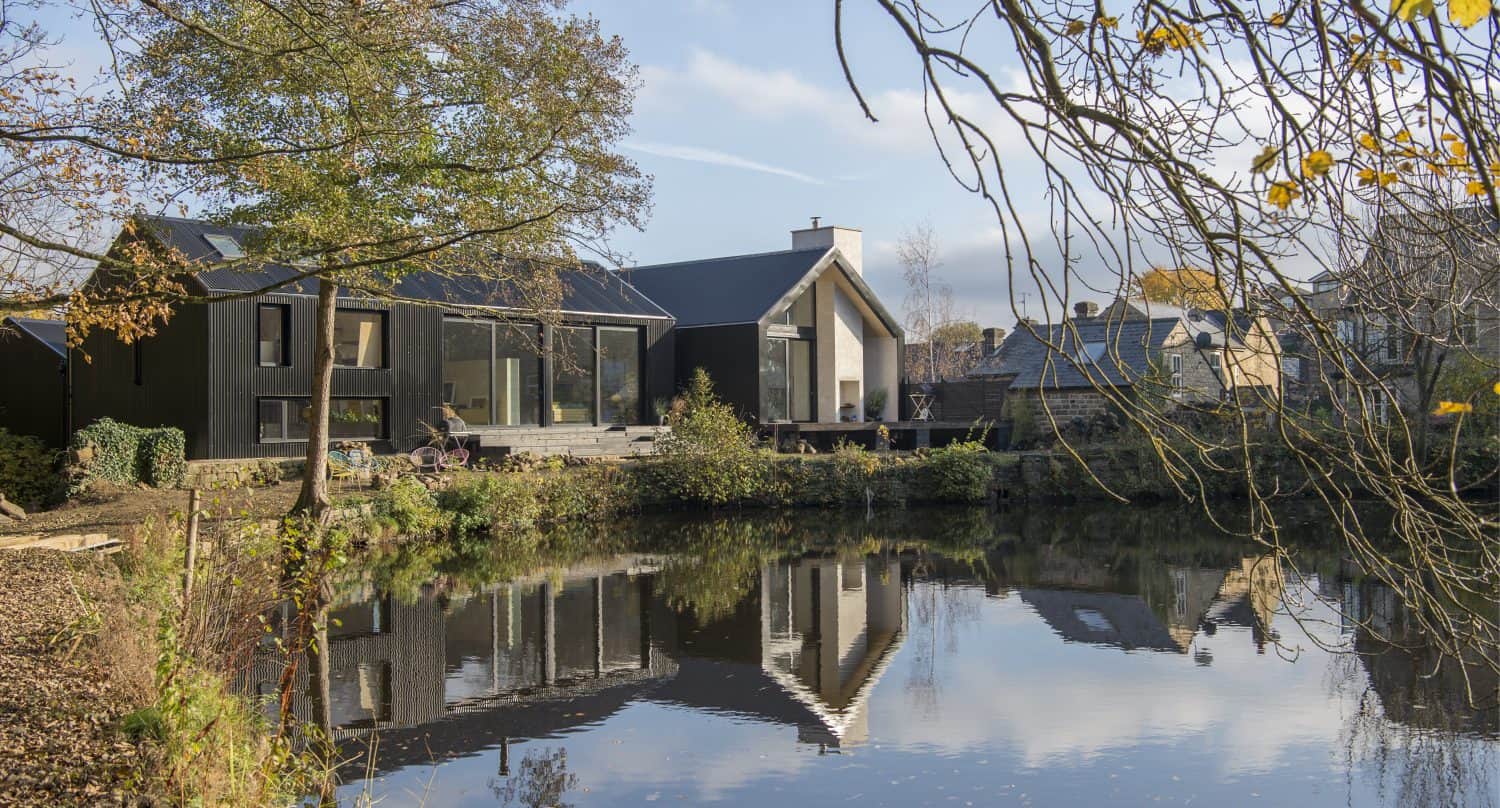 Modern house with large windows and glass roofing sits beside a calm pond, reflecting the building and autumn trees. The sky is clear, and other houses are visible in the background.