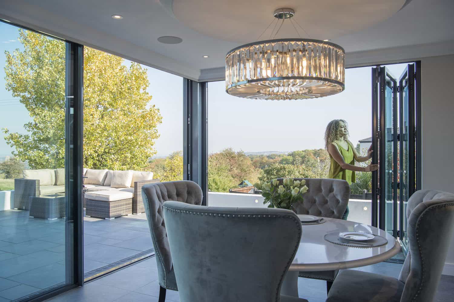 A woman stands by open sliding doors leading from a modern dining area to a spacious outdoor patio with seating, surrounded by trees and a scenic view. The room features elegant chairs and a chandelier.