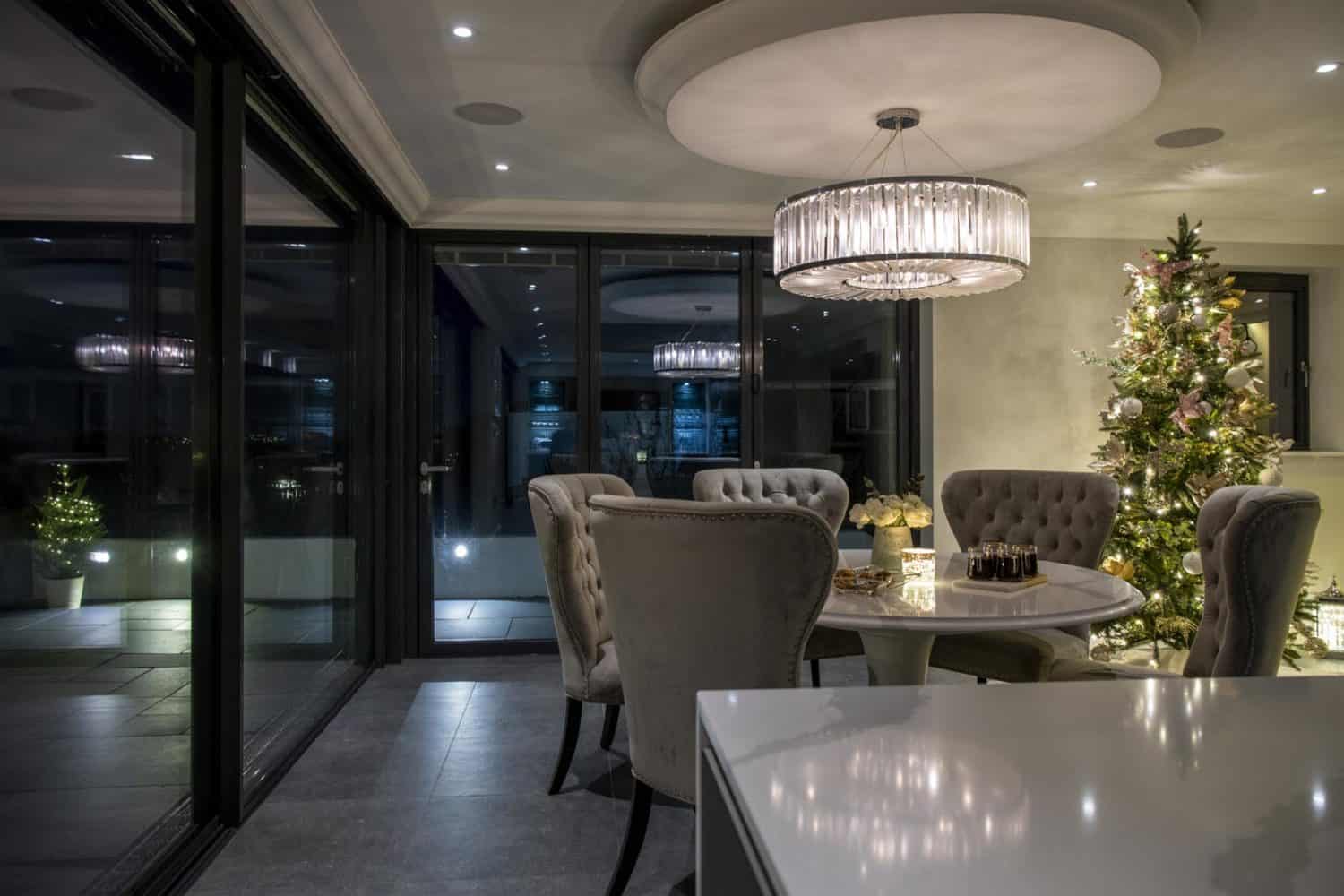 A modern dining room with a round table and tufted chairs, illuminated by a circular chandelier. A decorated Christmas tree stands in the corner, and large bifold doors open to a patio adorned with outdoor lights.