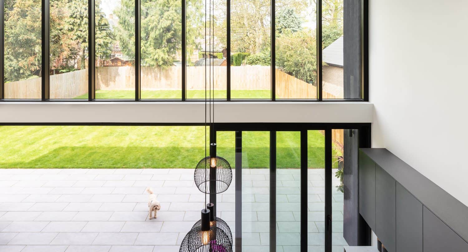Modern living space with large, black-framed windows and bifold doors overlooking a sunny backyard with green grass; a small dog stands outside on a tiled patio. Decorative pendant lights hang from the ceiling.