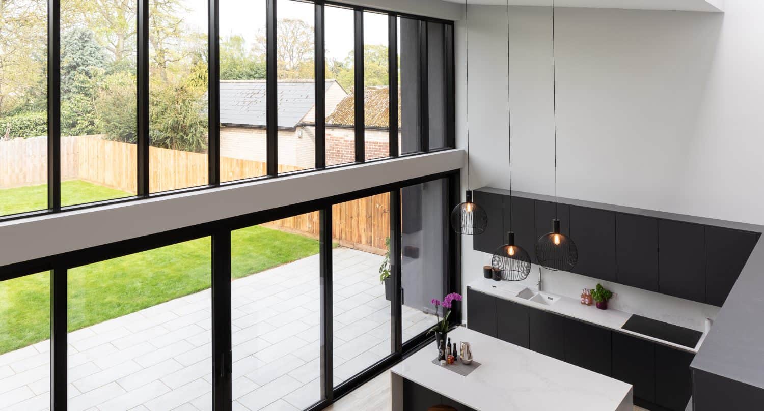 Modern kitchen with dark cabinets and white countertops, large windows and bespoke glass solutions like sliding or bifold doors opening to a patio and garden, pendant lights above the island, and stools at the counter.